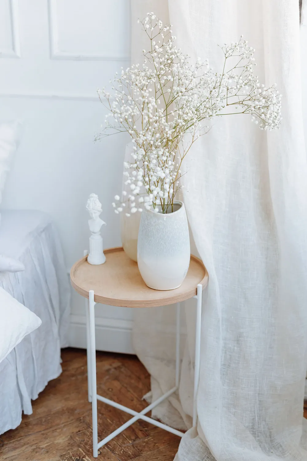 White ceramic vase with baby's breath on a light timber side table beside linen curtains