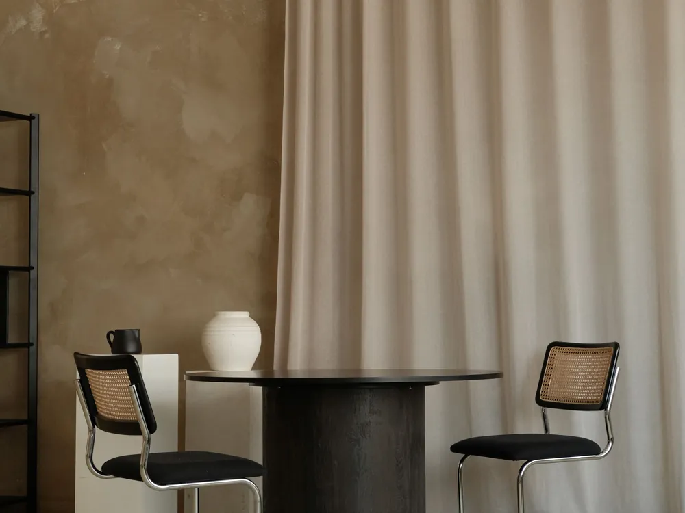 Round dark dining table with cane-back chairs against linen curtains